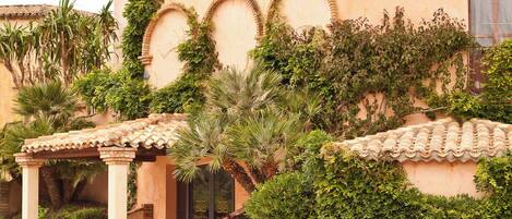 A building with arched windows and a tiled roof, surrounded by lush greenery and flowering plants.