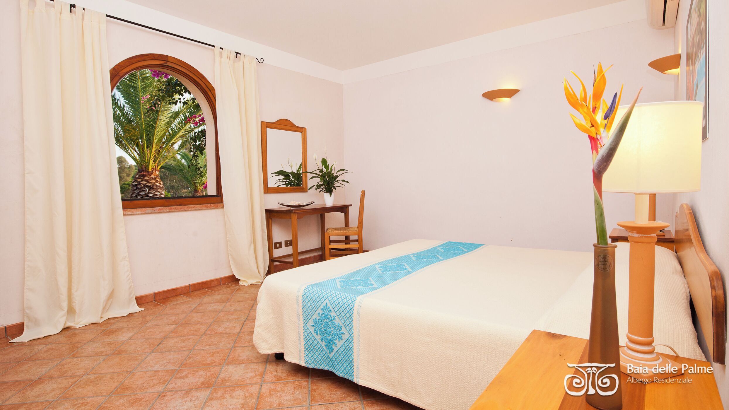 Ein Hotelzimmer mit Bett, Schreibtisch, Stuhl, einem Fenster mit Vorhängen, einem Spiegel, einer Pflanze und einer Lampe.