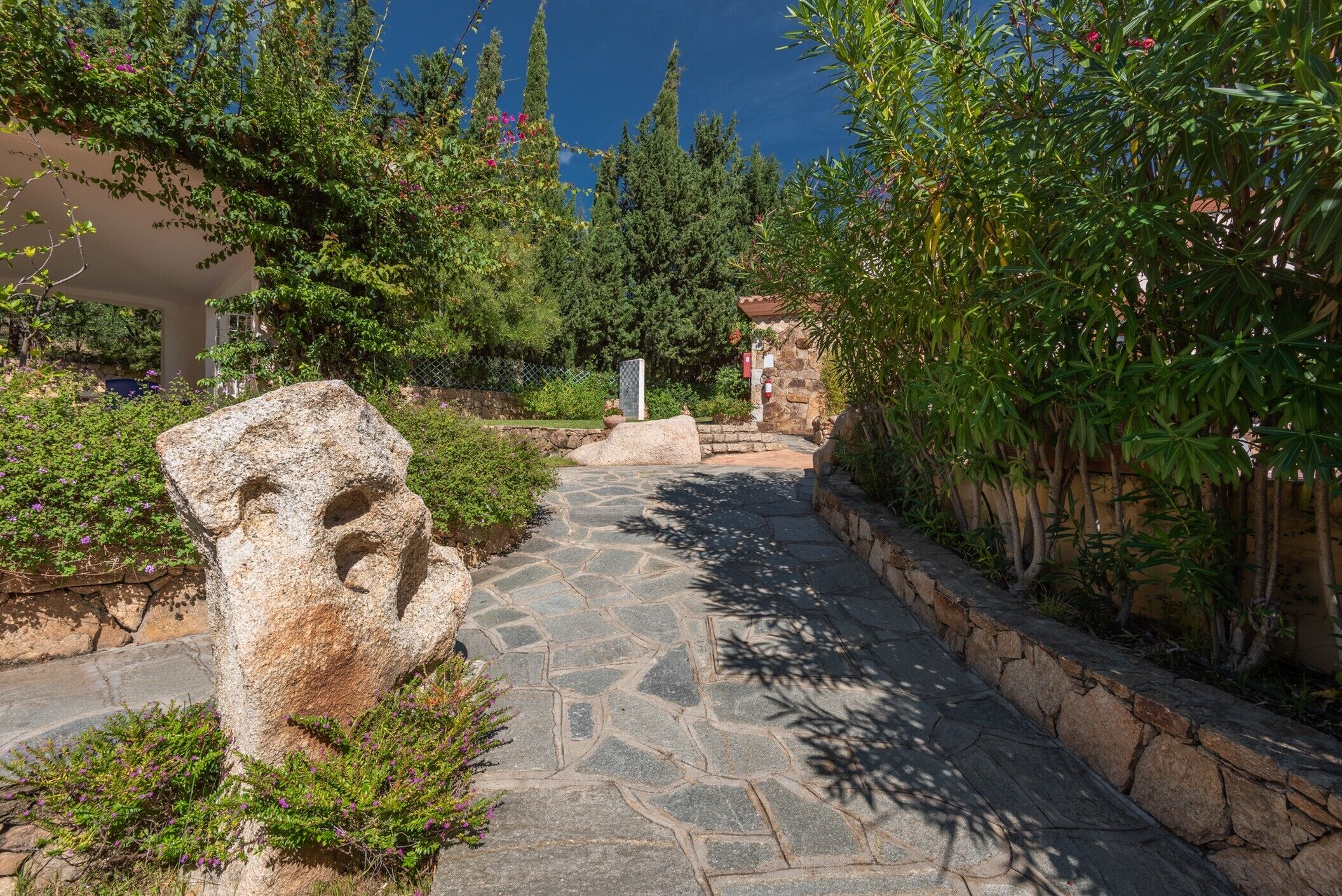 A stone pathway leading through a garden with a large rock sculpture and a stone wall on one side.