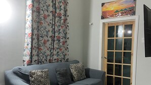 A living room with a grey sofa, patterned cushions, a grey rug, a wooden door with glass panels, and a floral curtain.