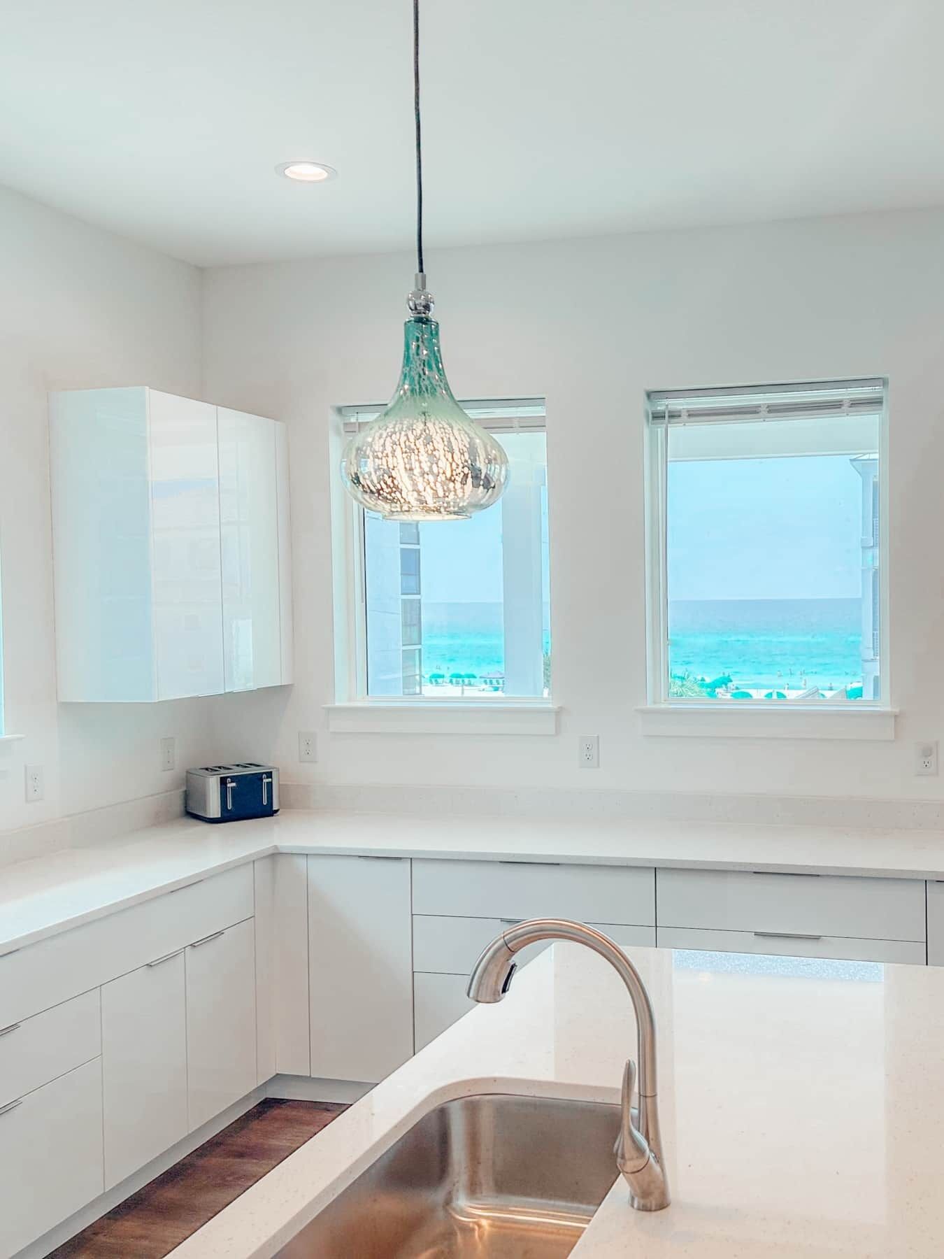 A modern kitchen with white cabinets, a stainless steel sink, and a view of the ocean through two windows.