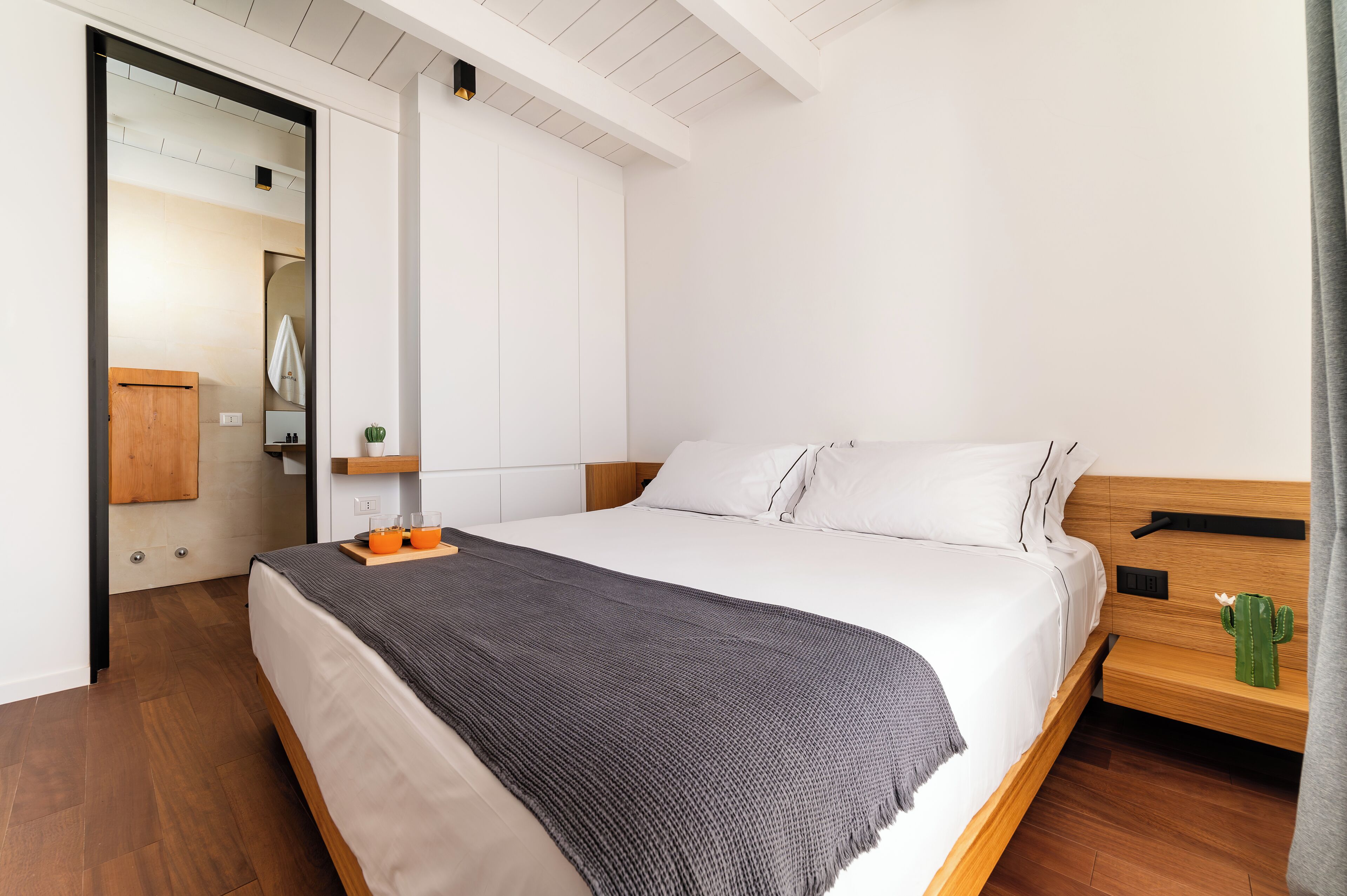 Un letto rifatto con un copriletto grigio, una testiera in legno e un comodino con un cactus.