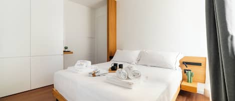 Una cama bien tendida con sábanas blancas, un cabecero de madera y una mesita de noche con una planta verde y dos tazas.