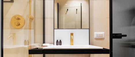 Un baño moderno con ducha envuelta en vidrio, un lavamanos blanco con grifo dorado y un espejo grande.