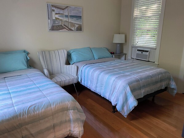 A bedroom with two beds, a nightstand with a lamp, a radiator, and a painting of a beach scene. - Cottage at Summer Village Gated Waterfront Resort Community-20 Miles from Boston (Westford)