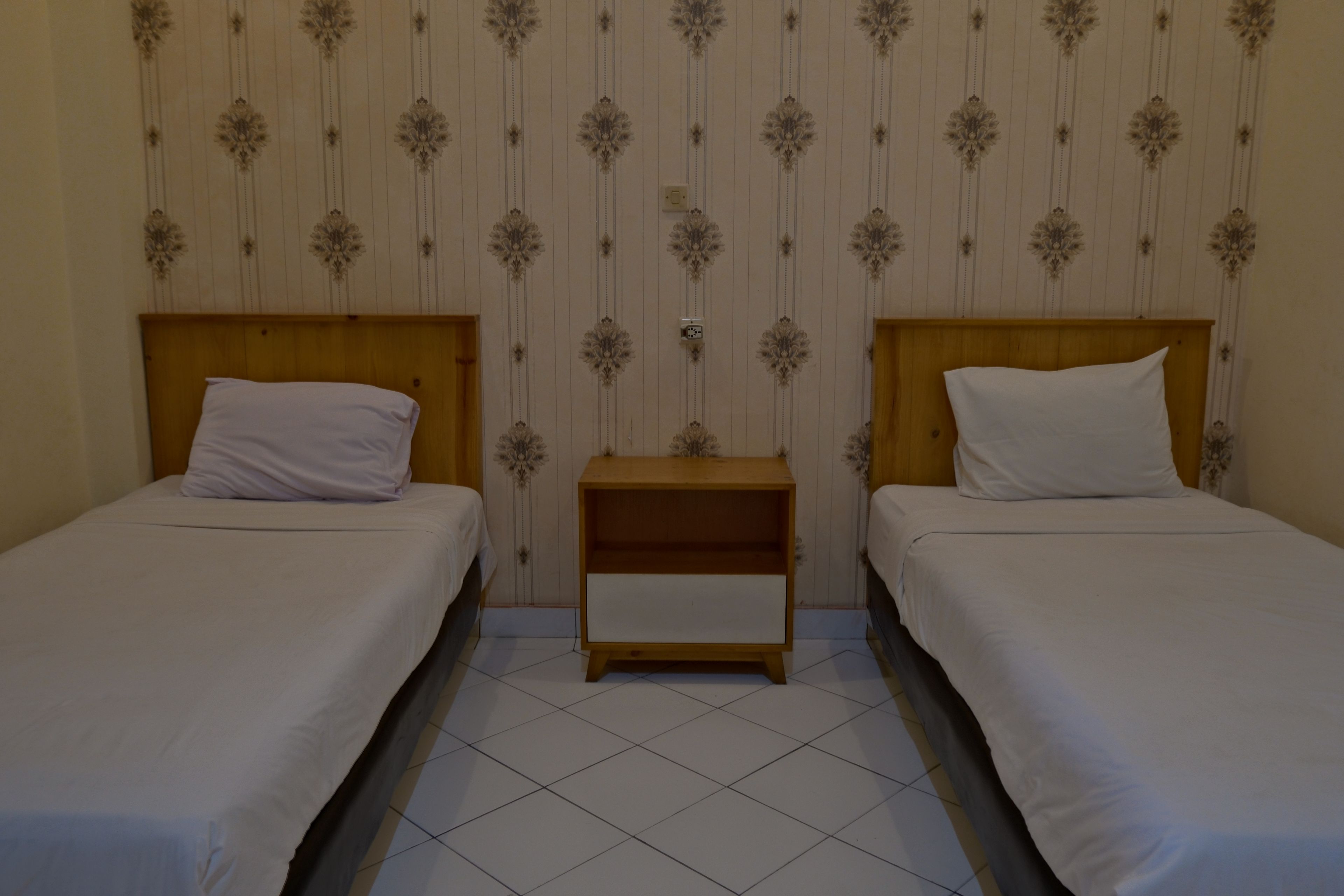 Une chambre avec deux lits simples, chacun doté d’une tête de lit en bois et de linge de lit blanc. Une petite table de chevet en bois est placée entre les lits. Les murs sont recouverts d’un papier peint décoratif à motifs verticaux.