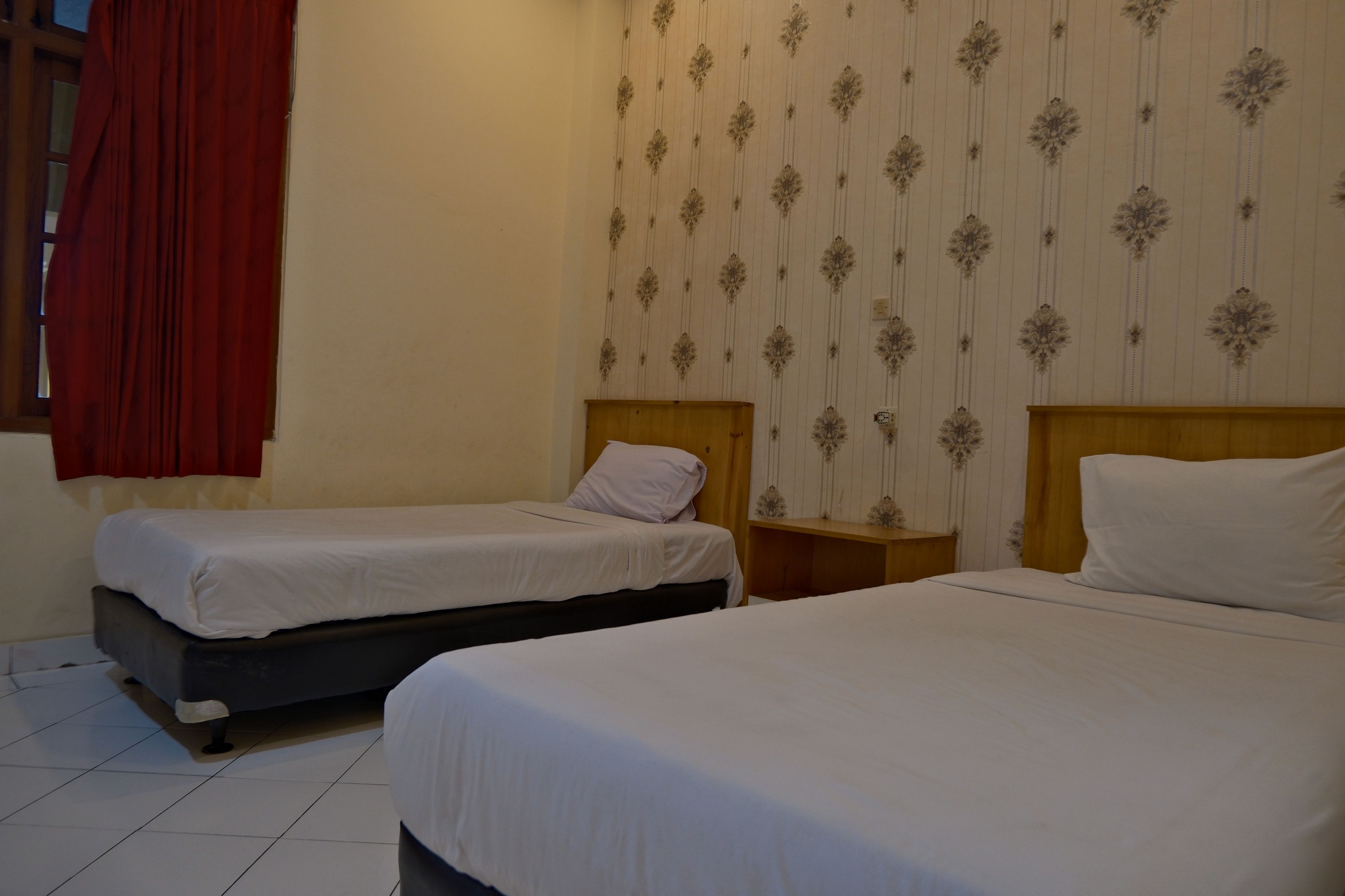 A hotel room with two single beds, a wooden nightstand, a red curtain, and a patterned wallpaper.