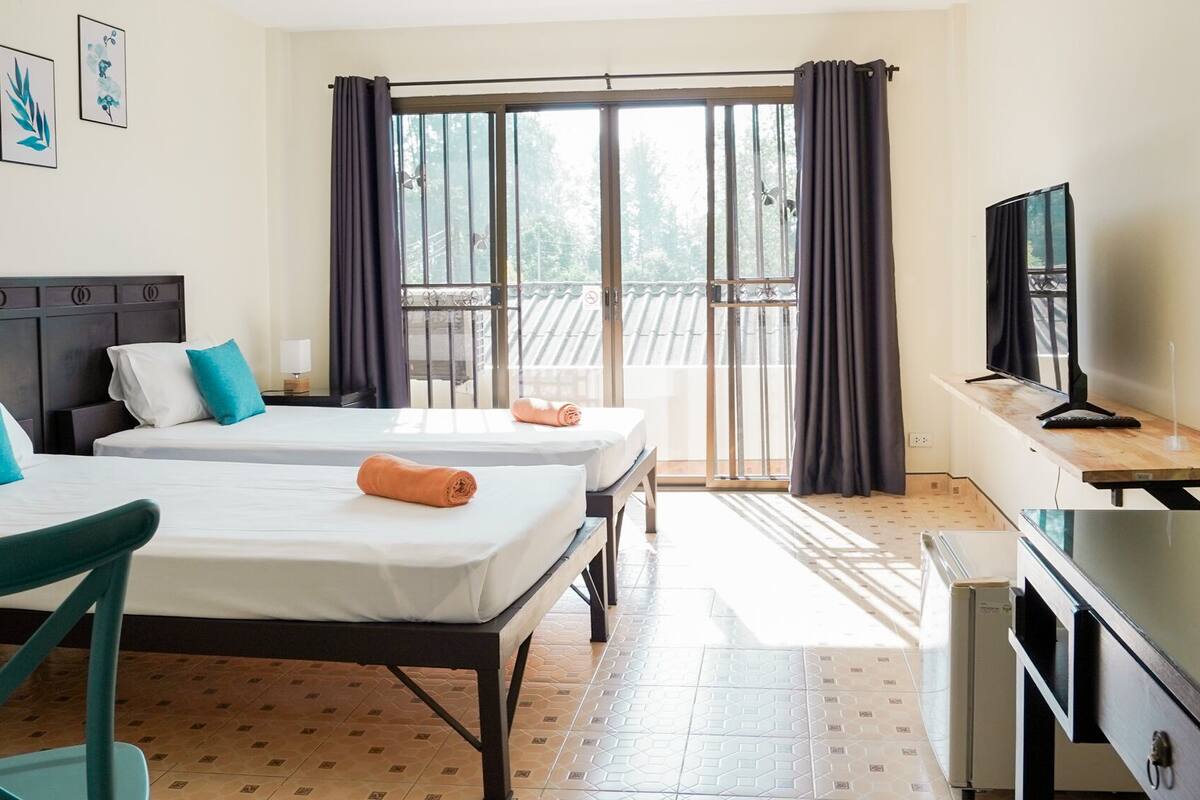 A hotel room with two beds, a TV, a desk, and a balcony with curtains.