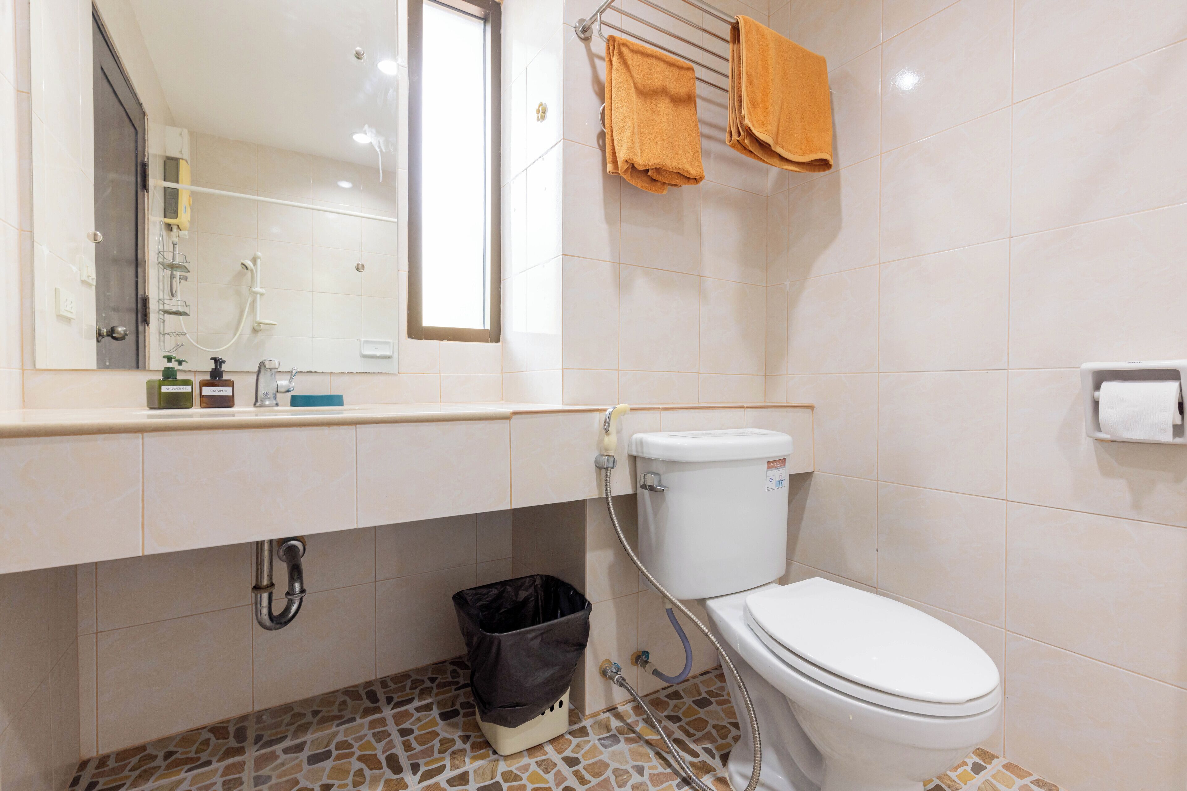 Un baño con inodoro, lavamanos y ducha. Hay toallas colgadas en un perchero y un cubo de basura debajo del lavamanos.