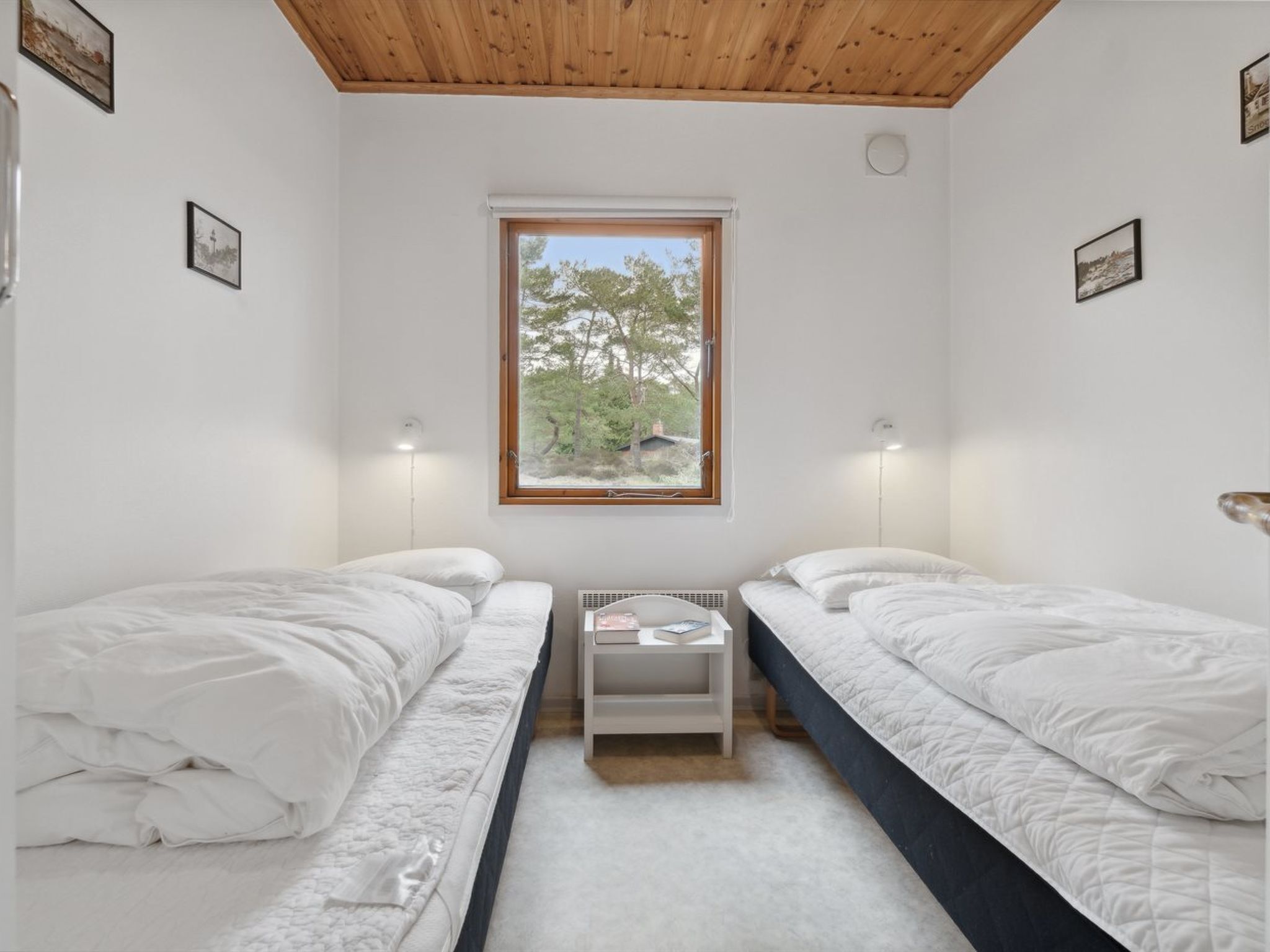 A bedroom with two single beds, a window with a view of trees, and a white bedside table with a lamp.