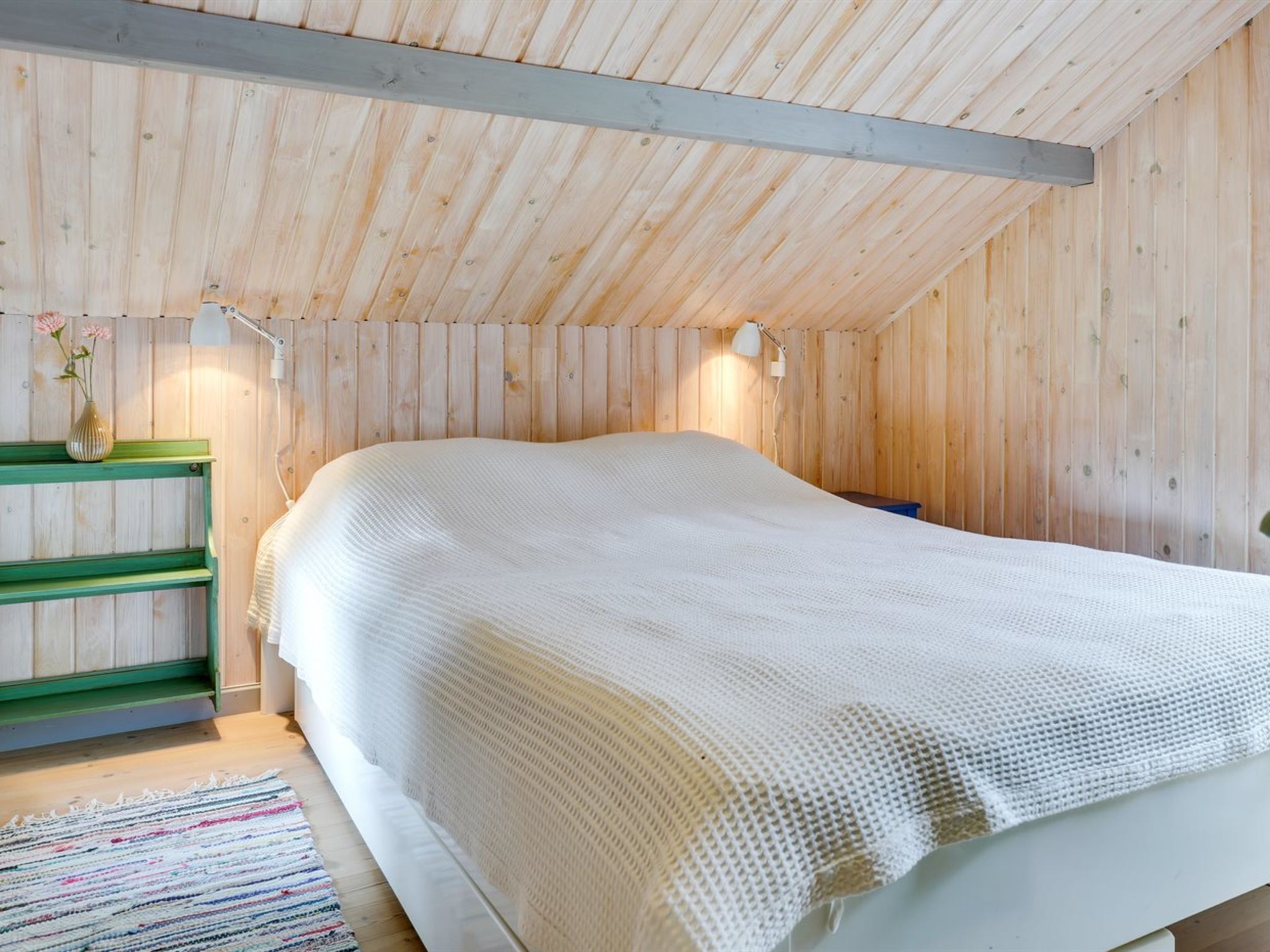 A room with a wooden ceiling, a bed with a white bedspread, a green bench, and a small table with a vase of flowers.