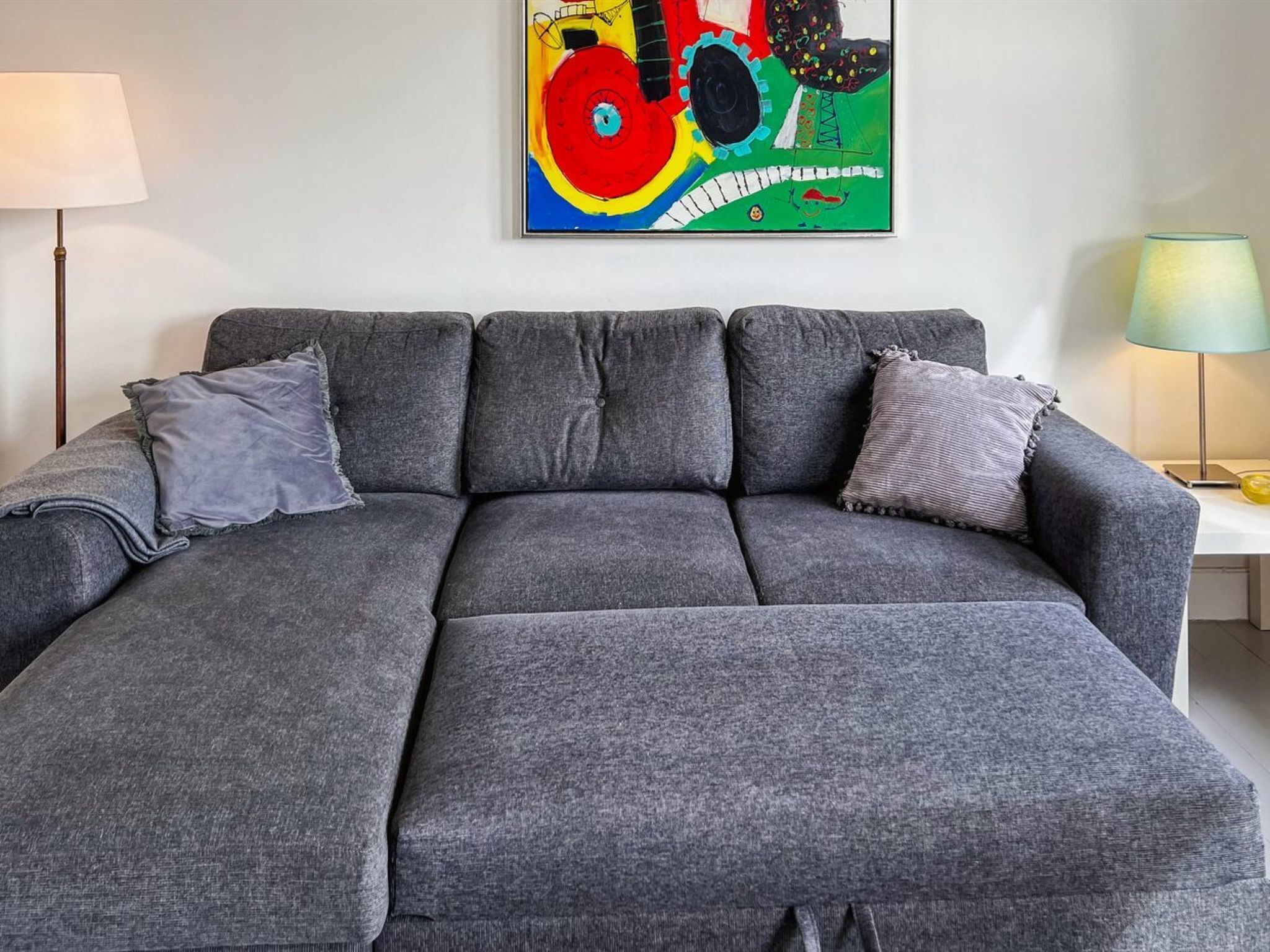 A grey sectional sofa with two grey cushions, a colorful abstract painting on the wall, a floor lamp, and a small table with a lamp.