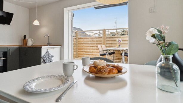 A modern kitchen with a dining area, featuring a large window, a dining table, and a breakfast setting with bread and a vase of flowers. - "Kiana" - 250m from the sea by Interhome (Nexø)
