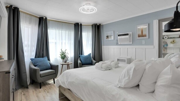 A modern hotel room with a large bed, a grey armchair, a small side table, and a window with curtains. - Capitola Village Hideaway Bungalow - Pet friendly! (Capitola)