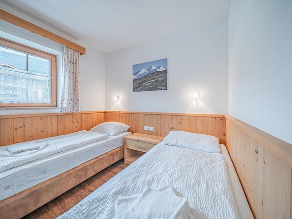Una habitación con dos camas individuales, cabeceros de madera, una ventana con cortinas, una lámpara de pared y un cuadro de paisaje montañoso.