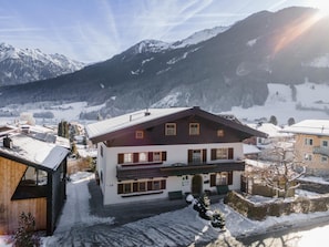 A two-story chalet with a balcony, surrounded by snow-covered mountains and a clear sky.