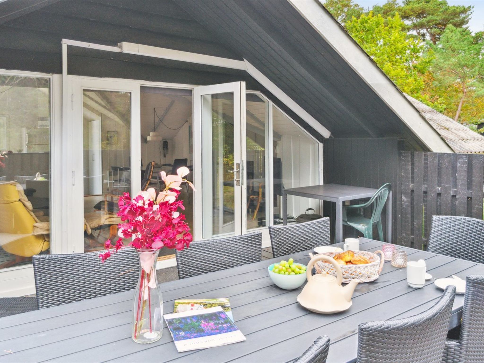 Ein Essbereich im Freien mit einem Tisch für zwei Personen, einer Vase mit rosa Blumen und Blick ins Haus durch große Glasüren.