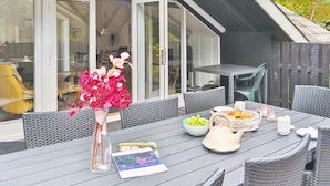 Un espace de restauration en plein air avec une table dressée pour deux personnes, un vase rempli de fleurs roses et une vue sur l’intérieur de la maison à travers de grandes portes vitrées.