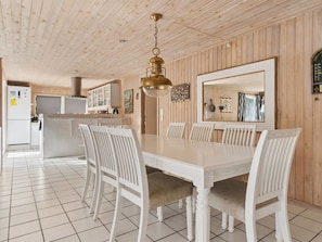 A dining area with a large table and chairs, a hanging light fixture, and a kitchen in the background.
