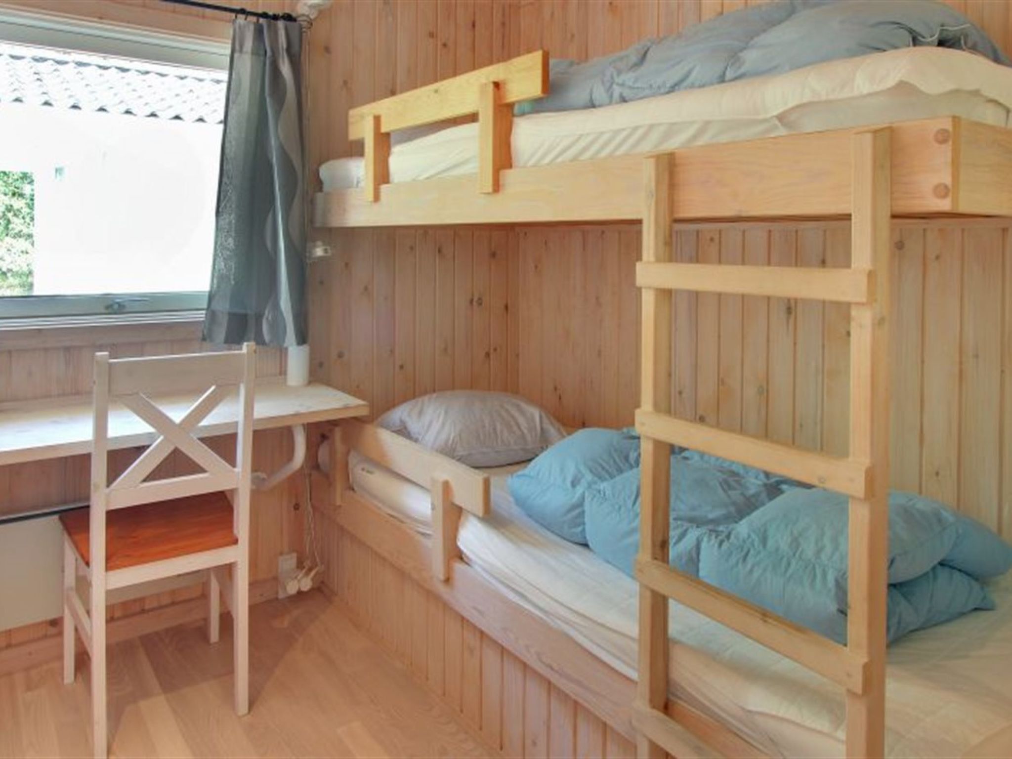 A bunk bed room with a desk and chair, wooden walls, and a window with curtains.
