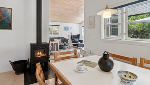 Una habitación acogedora con un estufa de leña, una mesa de comedor con sillas, un ventanal con vistas al verdor y una lámpara colgante.