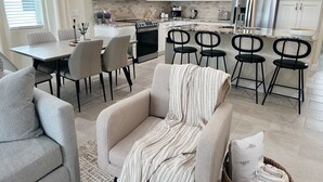 A modern kitchen with white cabinets, a stainless steel refrigerator, a microwave, and a coffee maker. There is a dining area with a table and chairs, and a living area with a sofa and a patterned throw blanket. - NEW Cozy Home in Historic Venice (Venice)