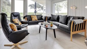 A modern living room with a black leather sofa, a black leather chair, and a wooden chair with a striped backrest. There is a round coffee table with a teapot and a small plant. The room has large windows with a view of trees and a grassy area. - "Botolphus" - 20m from the sea by Interhome (Nexø)