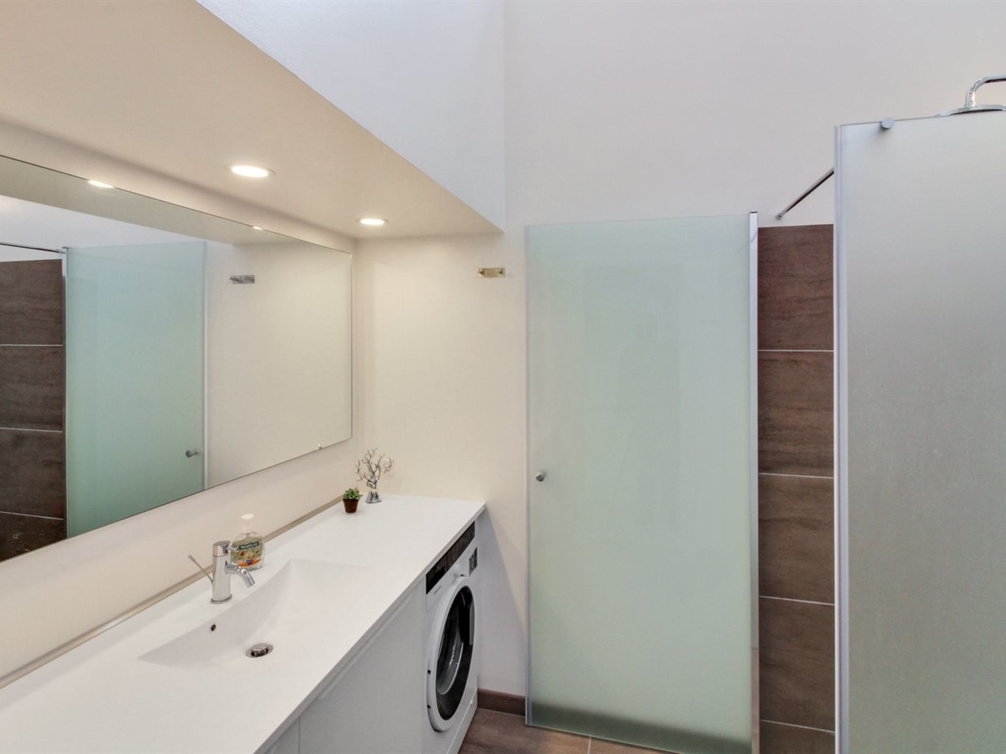A modern bathroom with a white sink, a mirror, a washing machine, and a frosted glass shower enclosure.