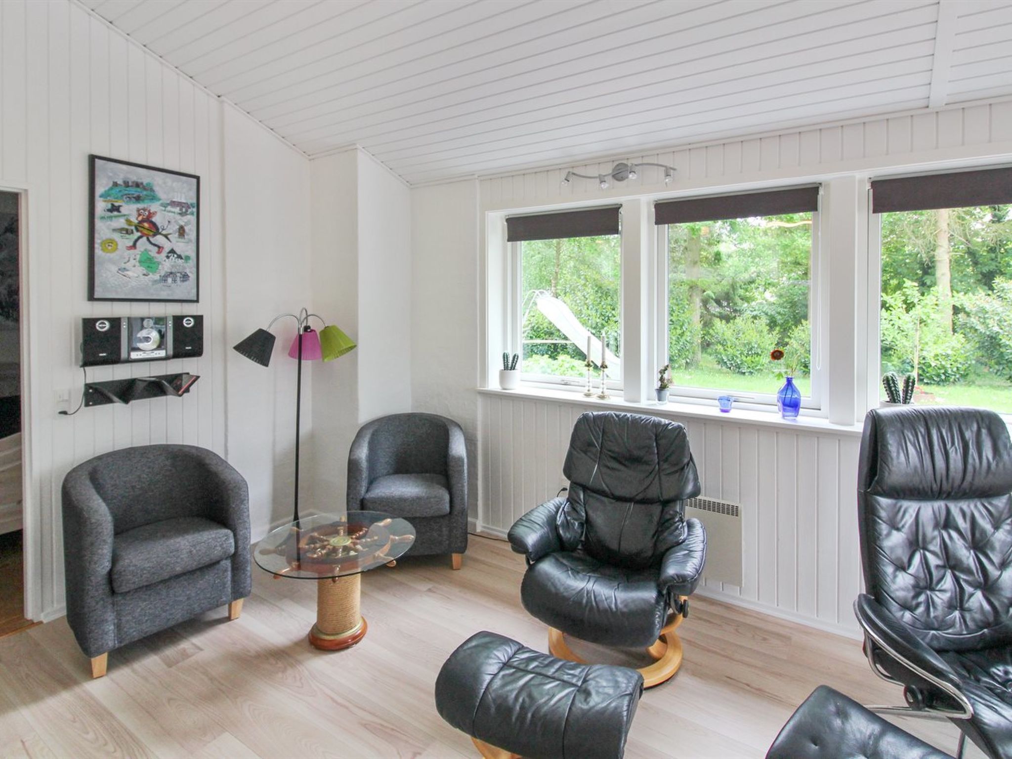 Une pièce comprenant deux fauteuils gris, un fauteuil en cuir noir, une table en verre et une fenêtre donnant sur un paysage verdoyant.