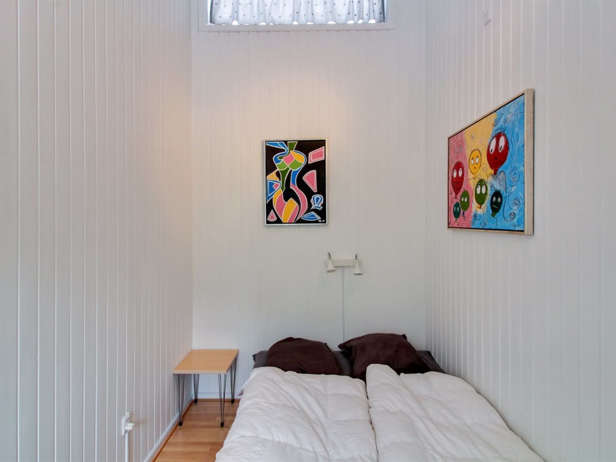 A bedroom with a bed, a small table, and two paintings on the wall.