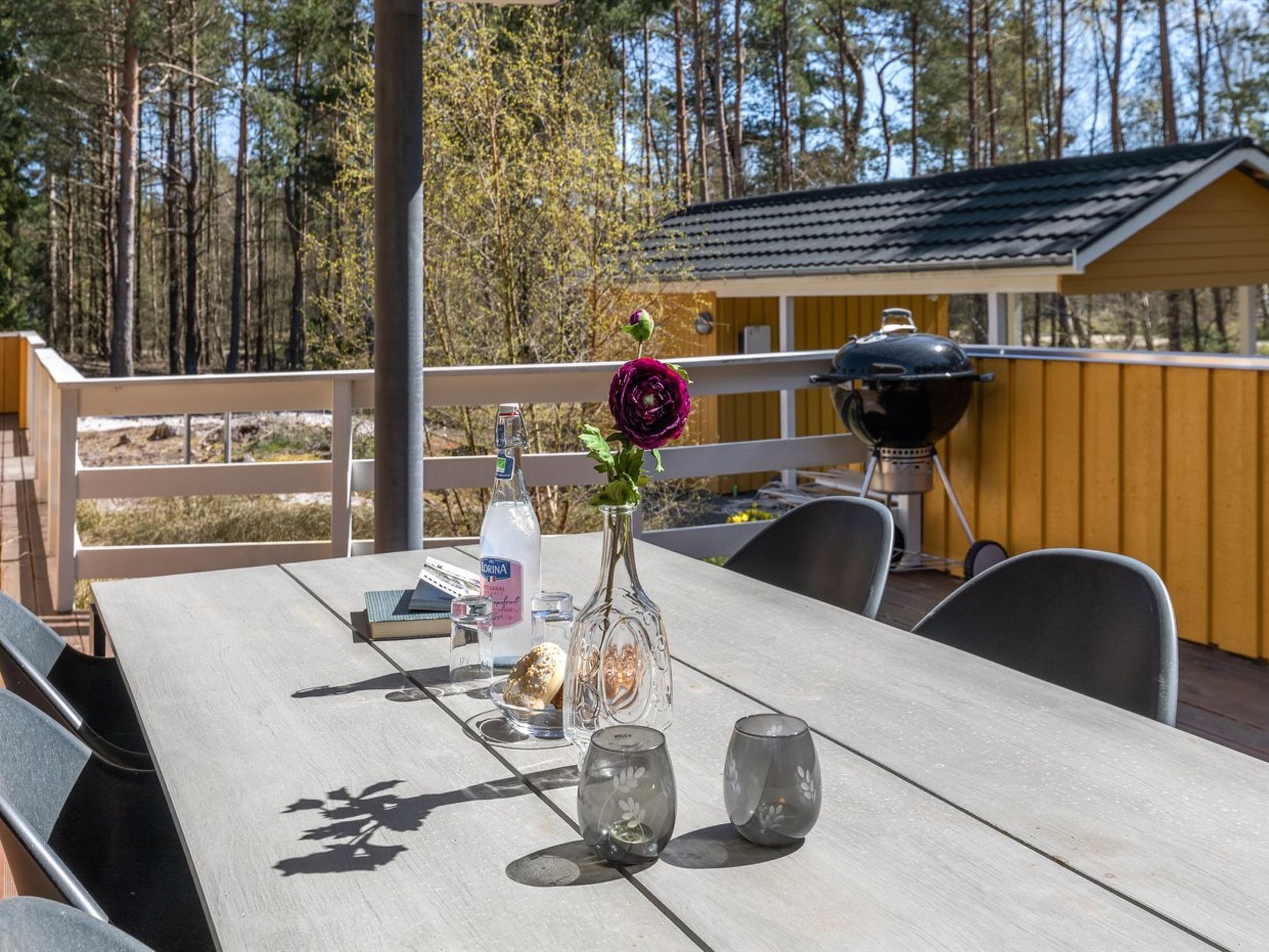 Una terraza de madera con una mesa preparada para dos, una botella de agua, un vaso y un jarrón con una flor.
