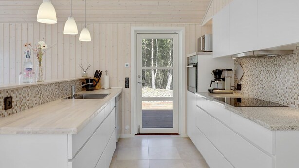 A modern kitchen with white cabinetry, a large island, hanging pendant lights, and a view through a glass door to an outdoor area. - "Arndis" - 600m from the sea by Interhome (Nexø)