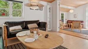 Uma sala de estar moderna com teto de madeira, um sofá de couro preto, uma mesa de jantar com pratos e xícaras, e vista para uma floresta através de grandes janelas.