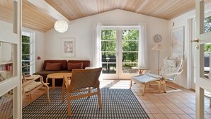 Um quarto aconchegante com teto de madeira, um sofá, uma mesa de jantar e uma cadeira. Há uma janela grande com vista para o verde lá fora.