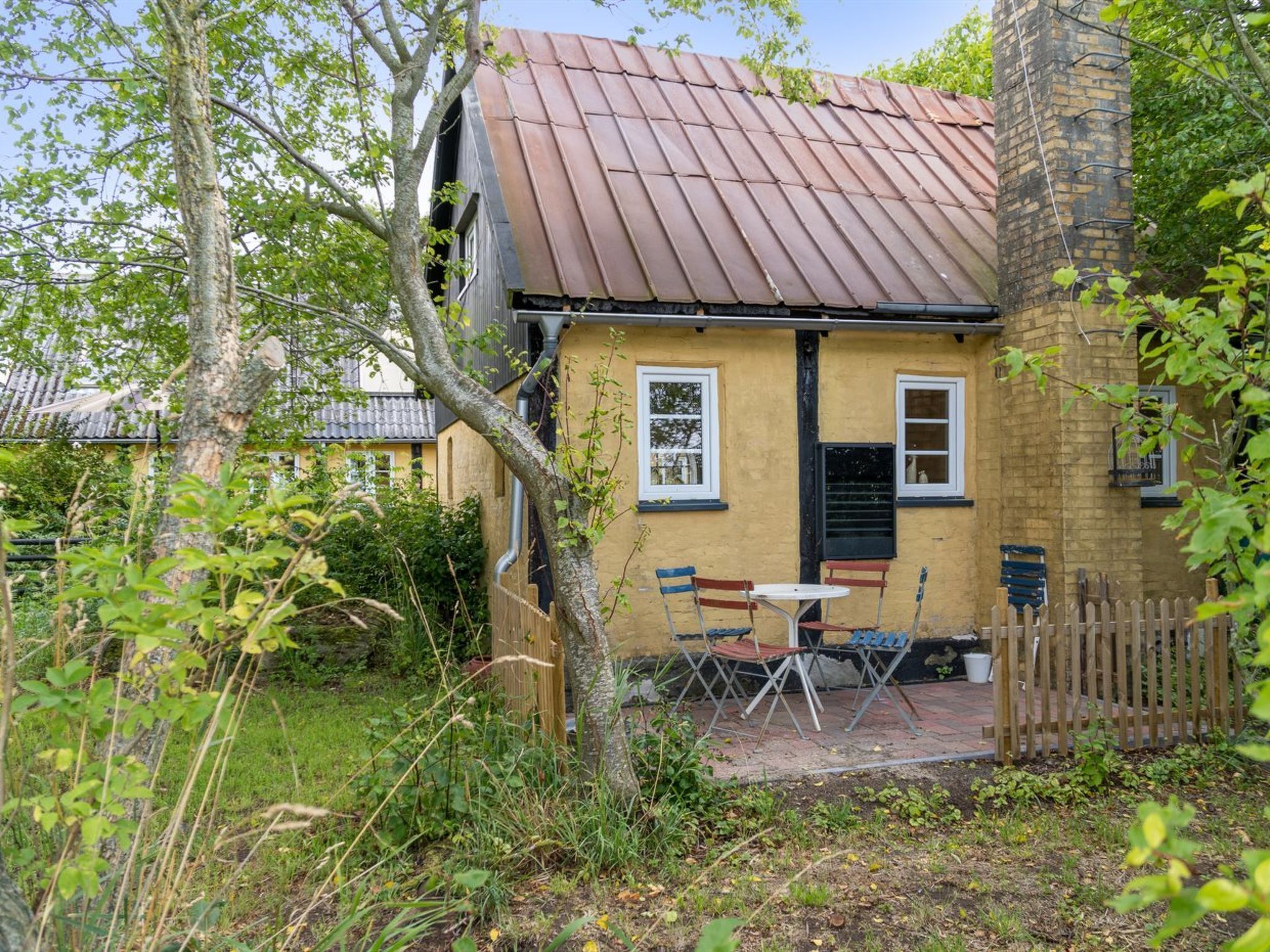 A small house with a yellow exterior, a brown roof, and a brick chimney. There is a table and two chairs outside.
