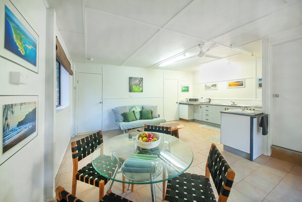A modern kitchen with white cabinets, a built-in oven, and a sink. There is a dining area with a glass table and chairs, and a sofa in the background.