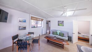 A modern living room with a glass dining table, wooden chairs, a sofa, and a ceiling fan.