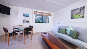 A living room with a wooden bench, a sofa with green cushions, a dining table with chairs, and a TV mounted on the wall.