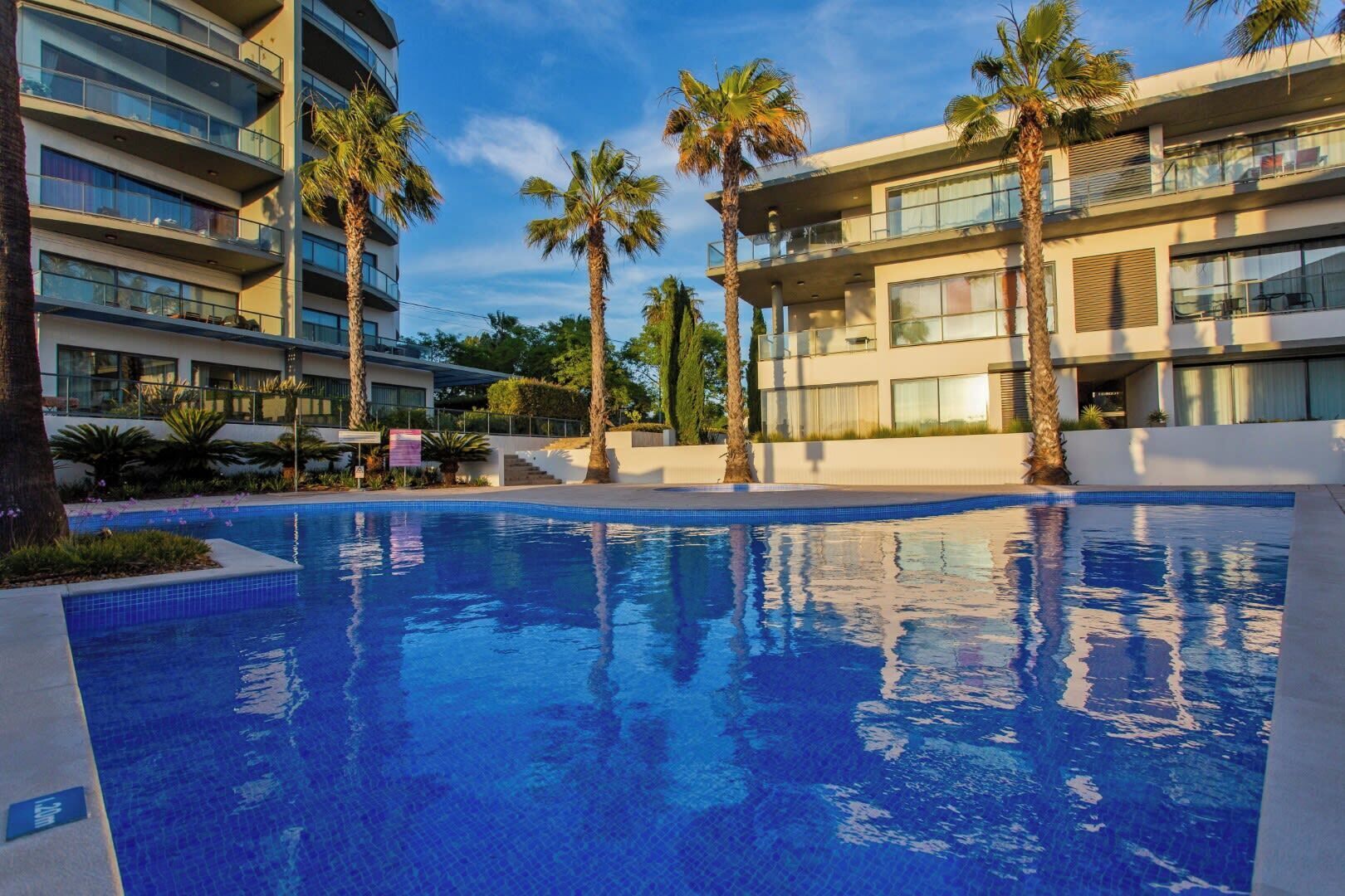 Um hotel moderno com piscina rodeada por palmeiras e um céu azul límpido.