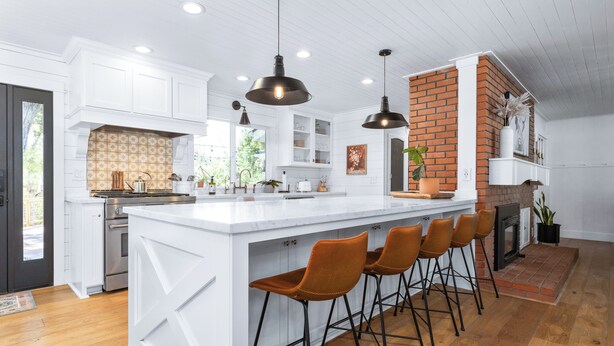 Uma cozinha moderna com armários brancos, uma ilha grande com banquetas, uma parede de destaque em tijolo e iluminação pendente.