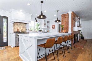 A modern kitchen with white cabinetry, a large island with bar stools, a brick accent wall, and pendant lighting. - Amazing! Wild Oaks Retreat. Pool, Spa, Game Room, EV Charger, Wine Country. (Atascadero)