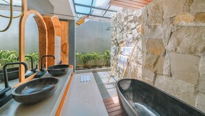 A modern bathroom with a stone wall, a skylight, and two large tubs. - Villa Asri -  Tropical 3BR Villa (Tabanan)