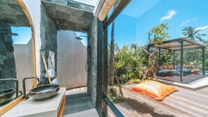 A modern bathroom with a stone archway, a wooden bench, and a view of a wooden deck and tropical plants outside. - Villa Asri -  Tropical 3BR Villa (Tabanan)