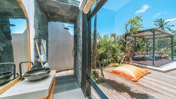 A modern bathroom with a stone archway, a wooden bench, and a view of a wooden deck and tropical plants outside.