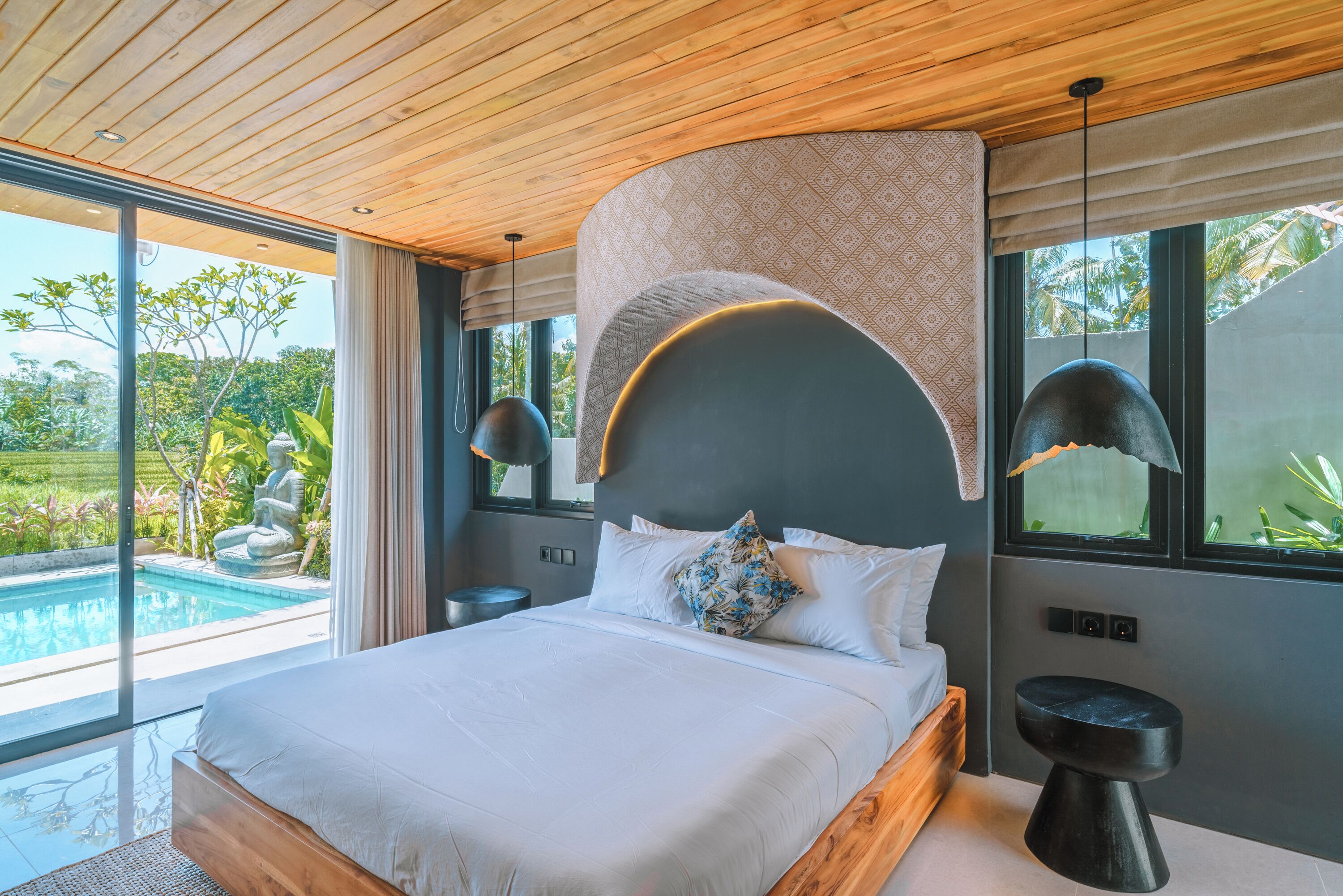 A modern bedroom with a large bed, a view of a swimming pool, and a wooden ceiling.