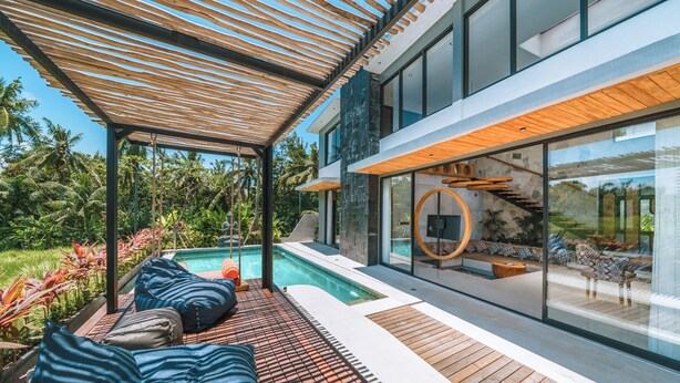 A modern outdoor pool area with wooden decking, lounge chairs, and a glass-enclosed staircase. - Villa Asri -  Tropical 3BR Villa (Tabanan)