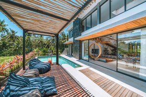 A modern outdoor pool area with wooden decking, lounge chairs, and a glass-enclosed staircase. - Villa Asri -  Tropical 3BR Villa (Tabanan)