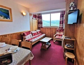 Ett mysigt rum med trägolv, en soffa med mönstrade kuddar, ett litet bord med en skål och en klocka, en stol med en hjärtformad kudde och en tv som är monterad på väggen.