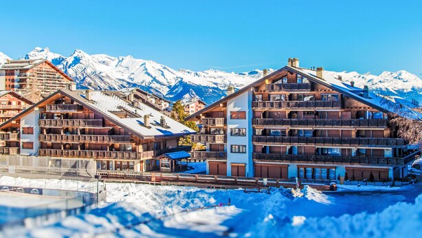 Ein Gebirgsort mit Holzbauten, schneebedeckten Bergen und blauem Himmel.