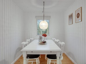 A white dining room with a large table, white chairs, a hanging light fixture, and a window with a view of trees.